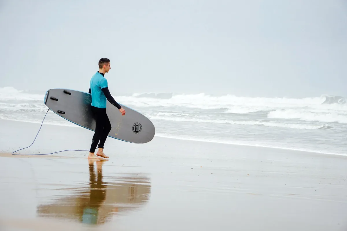 Student walking along Nazaré beach carrying a rental surfboard toward the water
